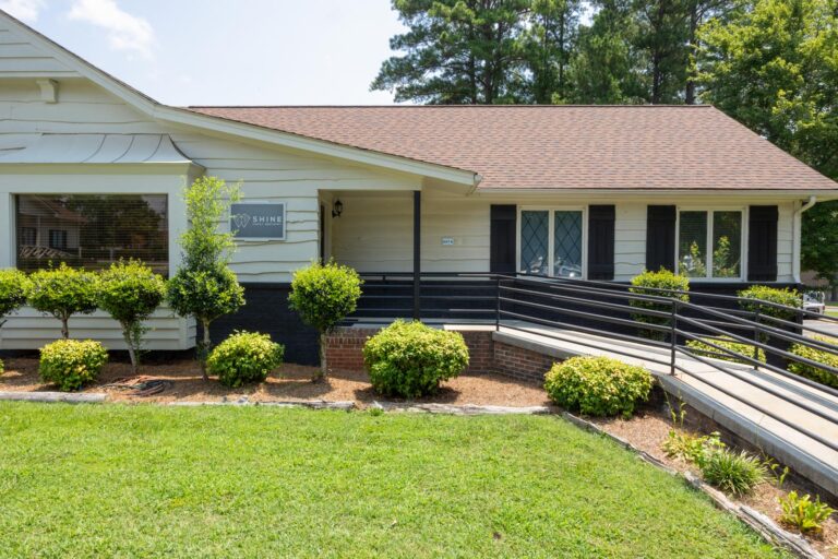 Exterior of Shine Family Dentistry featuring a white building with brown shingled roof, black shutters, and a ramp leading to the entrance; sign with “SHINE” and heart symbol mounted above the door, surrounded by trimmed bushes and a manicured lawn.