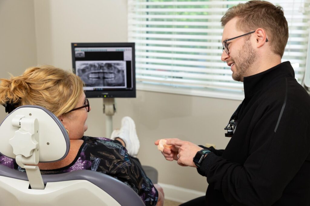 Dr. Shineholding a tooth model while explaining a dental X-ray displayed on a computer monitor to a seated patient with blonde hair and glasses; natural light enters through a window with partially open blinds.
