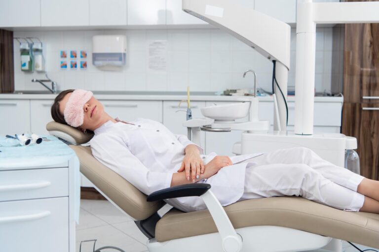 Person lying on a dental chair wearing a white coat and pink eye mask; surrounded by dental equipment, including overhead light, sink, cabinets, and wall posters in a well-lit clinic.